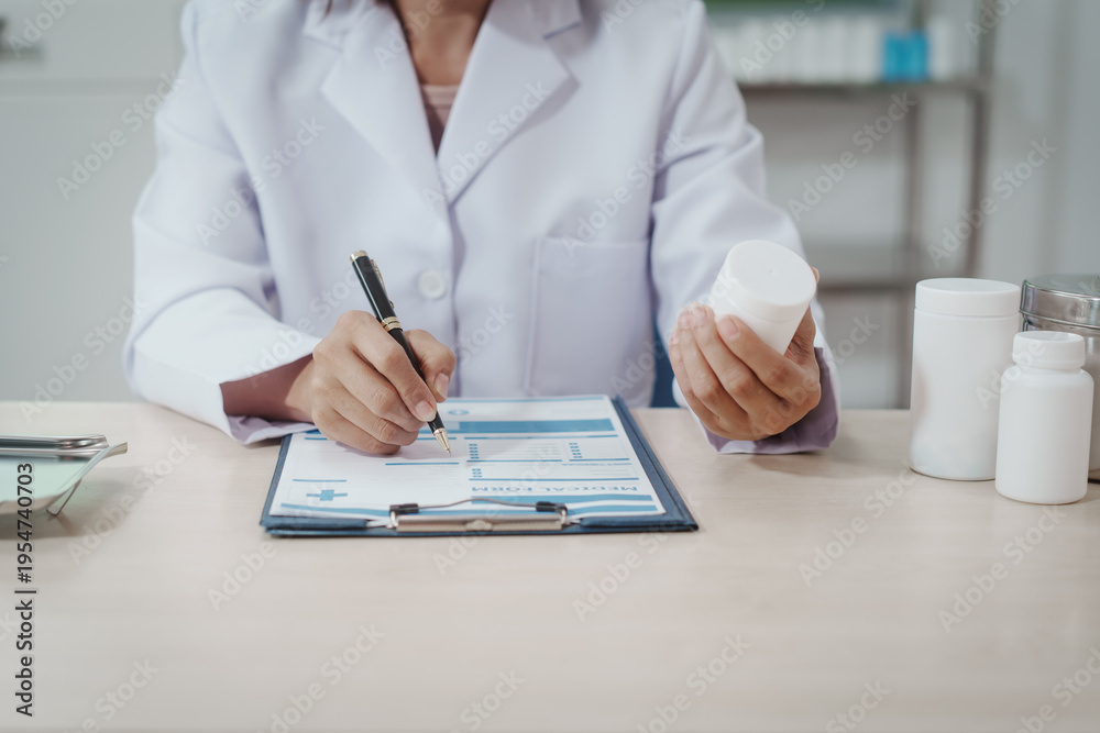 Fototapeta premium Doctor in white coat at desk writing on clipboard while holding medicine bottle, pharmacy containers on table, clinical office background