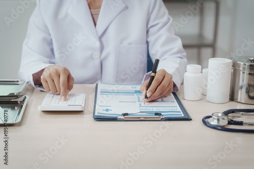 Doctor in white coat writing on clipboard beside pills, calculator and stethoscope on desk, medical consultation or prescription paperwork in clinic office.