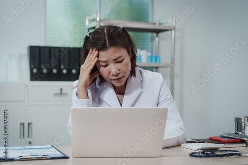 Stressed female doctor in white coat working on laptop at clinic desk, holding head with hand, paperwork and stethoscope nearby, healthcare burnout concept