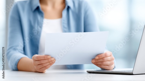 Woman in blue shirt holding a blank certificate paper in front of a laptop on a desk, with a blurred background suggesting an office or workspace environment