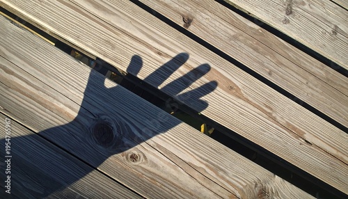 Close-up of hand's shadow on wooden deck, casting long shape between planks. Sunlight highlights textures and tones