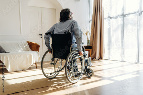 Slika na platnu Man in wheelchair facing large window in bright living room with wooden floor, s