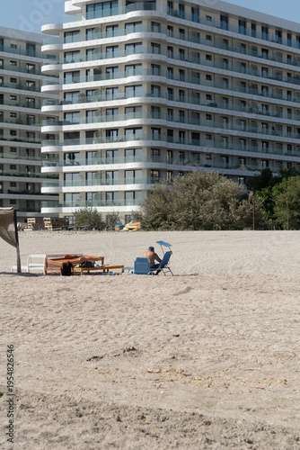 An unidentified man sunbathes on a beach on the Black Sea coast in Romania. View from behind. In the background are wooden pallets, vegetation, and a hotel building with glass balconies.