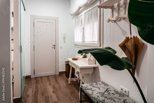 Modern home entryway with white wall, wooden floor, a decorative table, bench, coat rack, and a large plant