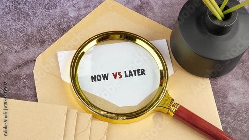 A golden magnifier artfully unveils the pivotal message NOW VS LATER on white paper. It rests on an envelope at a refined desk alongside a stylish black vase