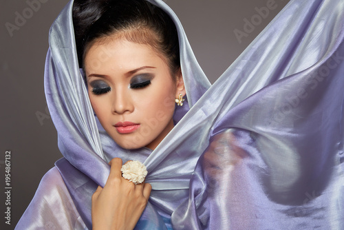 Woman with dark hair and smoky eyes, draped in a silky lavender scarf. She holds the fabric gently near her neck. Her expression is calm and slightly mysterious. The lighting highlights her features