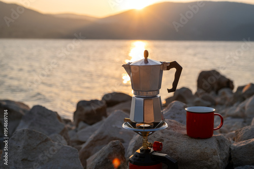 Red metal cup and moka pot on portable stove against peaceful lake and hills background at dusk, perfect scene for rustic travel, hiking break, nature lovers, coffee ritual