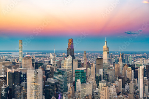 A high angle aerial view shows the dense skyline of Midtown Manhattan under a vibrant pink and orange sunset sky in New York.