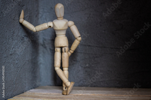 A wooden mannequin leans its arm against a gray wall, legs crossed. A confident, relaxed pose against the rough concrete texture. Stylish.