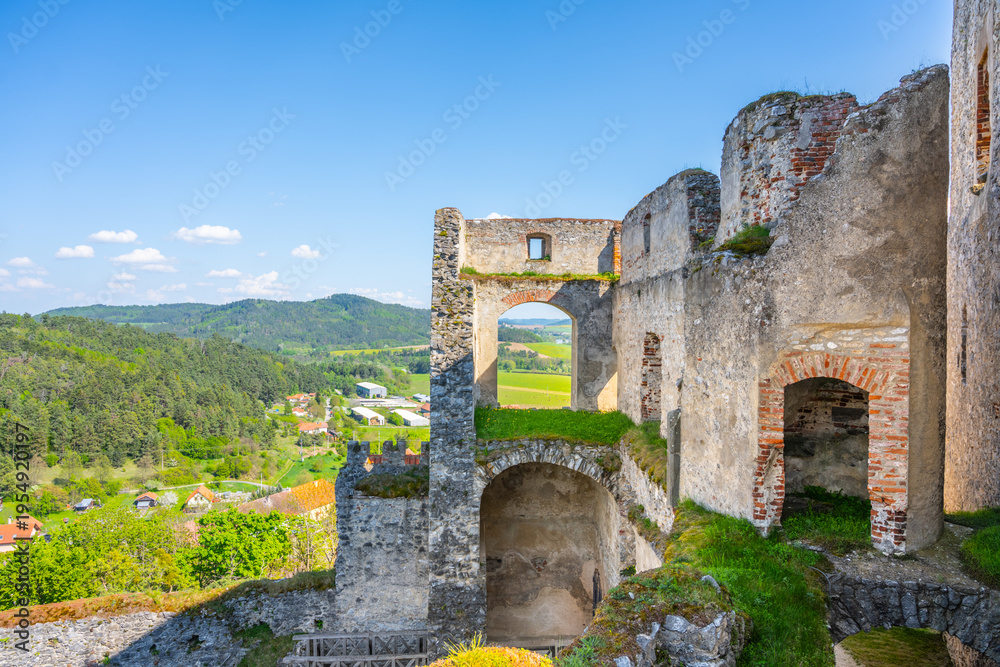 Obraz premium Rabi Castle stands as a large ruin in Bohemia. Visitors can see the stone walls and views of the village below. The sky is clear, and the landscape shows green hills and houses.