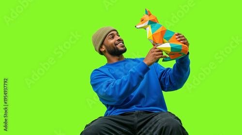 Smiling African American man holding a colorful geometric fox figurine. Happy male in blue sweatshirt looking at low poly animal toy on green screen background. Copy space for text