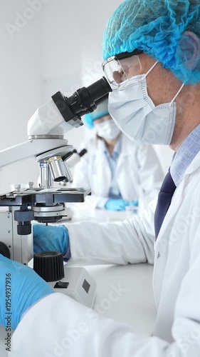 Senior male scientist wearing a protective mask, cap, and gloves, examining samples under a microscope in a modern laboratory. Medicine, healthcare and science concept