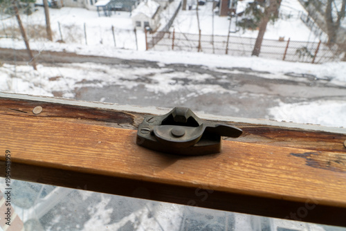 Quadro su tela Close-up of an old metal window lock on a rustic wooden sill, with a blurred, snowy winter landscape of fences and trees visible through the dirty glass, evoking a sense of cozy indoors