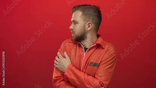 Tableau sur toile Man prisoner clutching his arms and bracing himself wearing an orange jumpsuit in a red studio; vulnerability