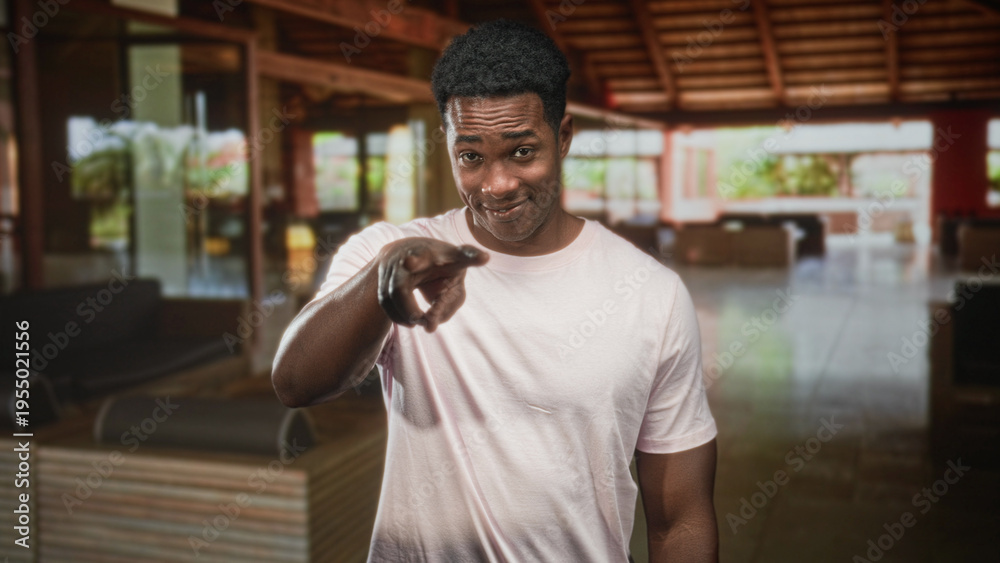 Obraz premium African american man in pale pink tshirt smiles at camera inside a hotel building lobby; friendly welcome.