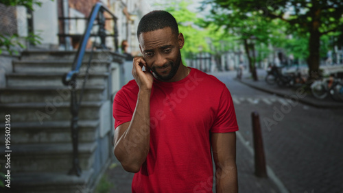 Man holding phone to ear with hand to face on city street near stone steps in red shirt; worry isolation.