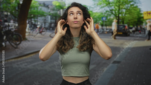 Hispanic young woman cups ears with hands amid bustling city street traffic and passing bicycles; noise frustration overwhelm.