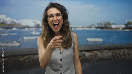 Konstfotografi Young hispanic woman wearing glasses and a light grey vest points finger by sunlit seaside street; playful