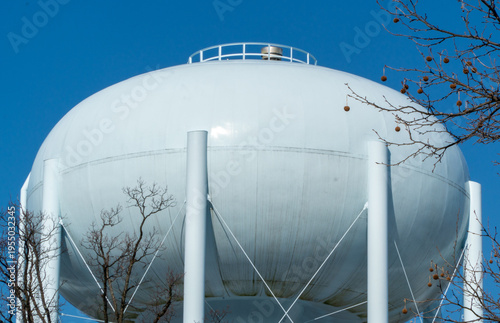 Φωτογραφία Giant white spherical water tower providing essential municipal water storage, standing tall against a vibrant clear blue sky, with bare winter tree branches framing the impressive utility structure
