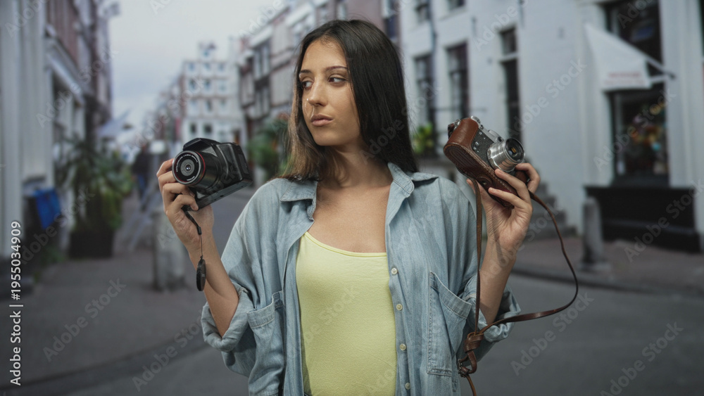 Fototapeta premium Woman holds two cameras in each hand on a city street while looking at the viewer, denim shirt visible; confidence exploration.