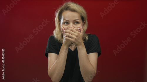Foto Blonde woman in black shirt with wide eyes, hands covering mouth and staring at camera in studio; surprise