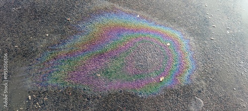 Close-up of an iridescent oil film on a textured grey asphalt surface. Vibrant rainbow colors highlight environmental pollution and chemical waste in an urban environment. Macro view.