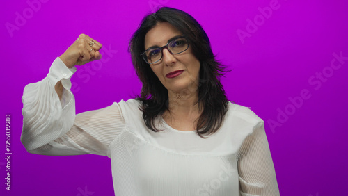 Cuadro en lienzo Middle aged hispanic woman wearing glasses firmly raises fist and flexes arm in purple studio; confidence