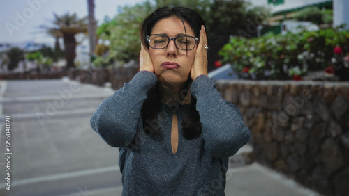 фотографія Middle age woman holds bare ears with hands on city street sidewalk against blurred stone wall and foliage; frustration