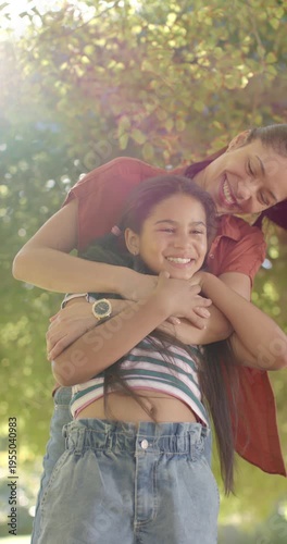 Pulling child close, mom behind daughter hugging, showing love under park tree, red tee, sun, watch