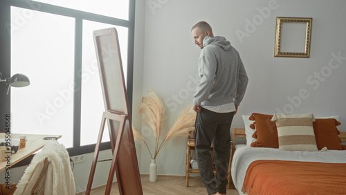 Wallpaper Mural Man pulls hoodie over head by bed and mirror, adjusts hood and checks face in full length mirror in a sunlit bedroom building; calm reflection. Torontodigital.ca