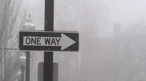 Fotografie One Way street sign with a prominent right arrow, standing firm in a dense, mystical fog, symbolizing a clear path or decision amidst an uncertain, hazy urban environment