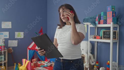 Photography Woman middleaged teacher holds clipboard and phone to ear, checking notes with red smartphone amid toys on shelves in building classroom; concern responsibility