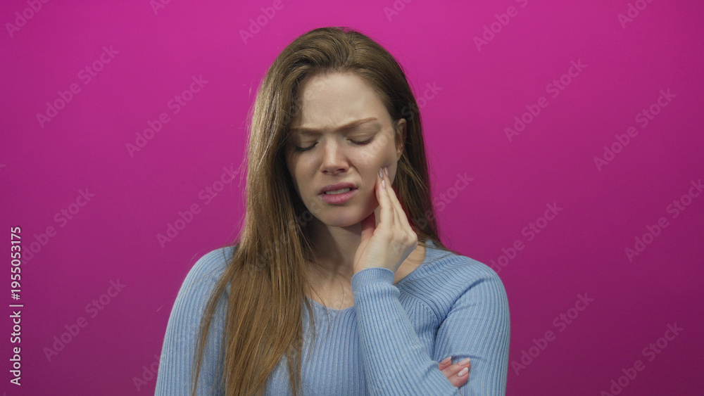 Fototapeta premium Young caucasian woman with pained expression hand on cheek in pink studio set; discomfort toothache.