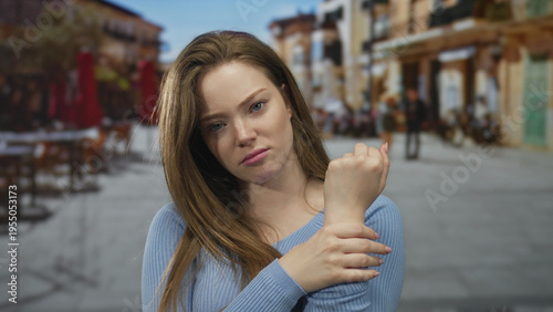 Foto Woman in blue sweater frowning holds wrist on busy sunlit street cafe terrace with outdoor tables; discomfort