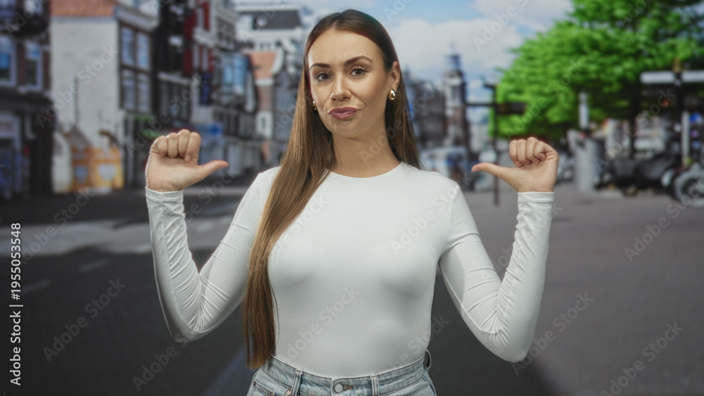 Fototapeta premium Woman with thumbs pointing to chest on a city street wearing white longsleeve top and jeans, long straight hair and gold studs, neutral smirk; confidence.