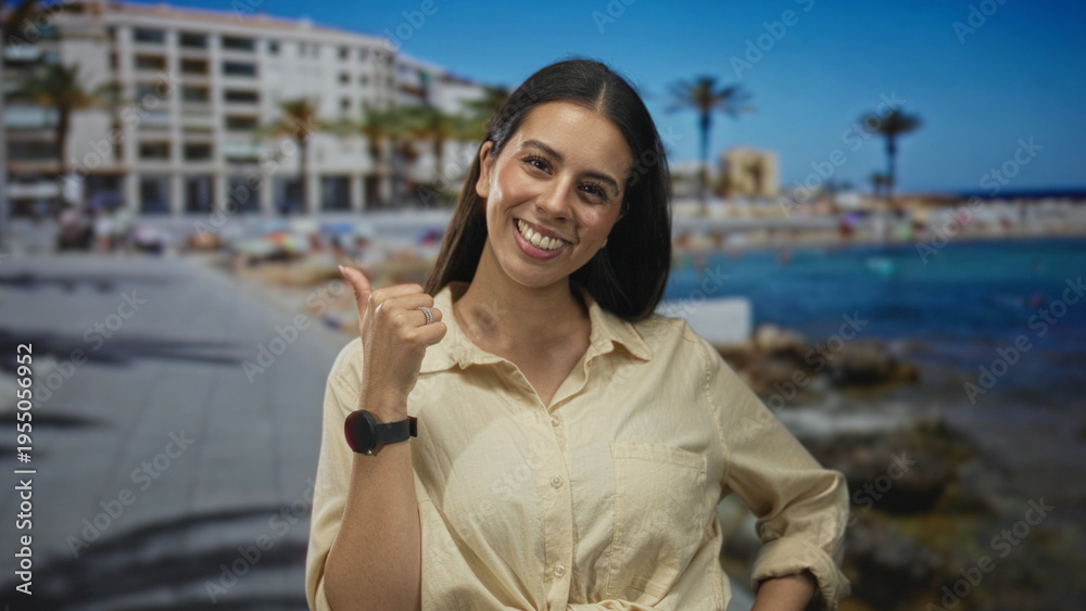 Fototapeta premium Young brunette woman smiling with thumb pointing to a seaside building on a sunny beach promenade near rocky shore; vacation joy relaxation.