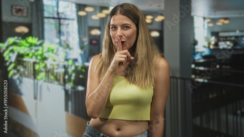 Wallpaper Mural Young woman in yellow crop top with index finger to lips making shushing gesture inside a bright cafe building near plants and pendant lights; playful secret. Torontodigital.ca