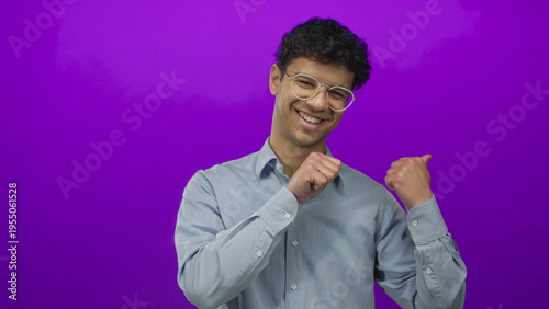 Foto Young hispanic man wearing glasses and a light blue shirt raises closed fists at shoulder level in bright purple studio; celebration joy