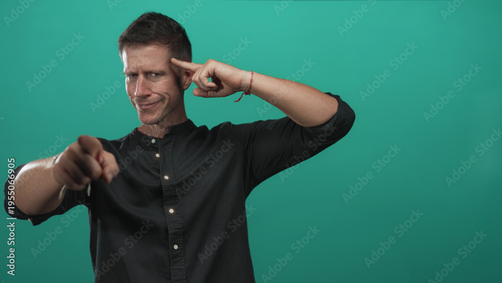 Fototapeta premium Man pointing finger at camera while touching temple with index finger in turquoise studio, wearing black shirt and smirking; confidence.