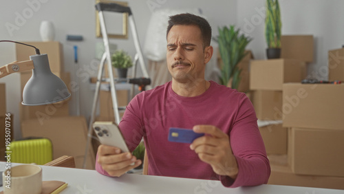 Bilde på lerret Young man with moustache holds smartphone and creditcard, frowning while checking a charge amid moving boxes in a building; financial frustration