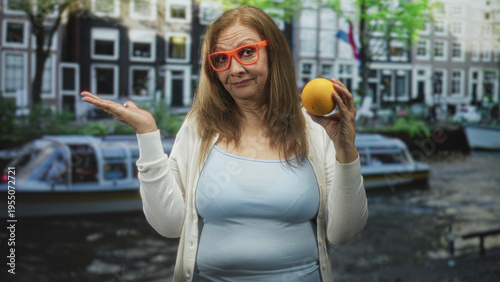 Canvas Print Woman wearing orange glasses and light blue top shrugs with palm up while holding an orange in a studio with canal backdrop; bemusement