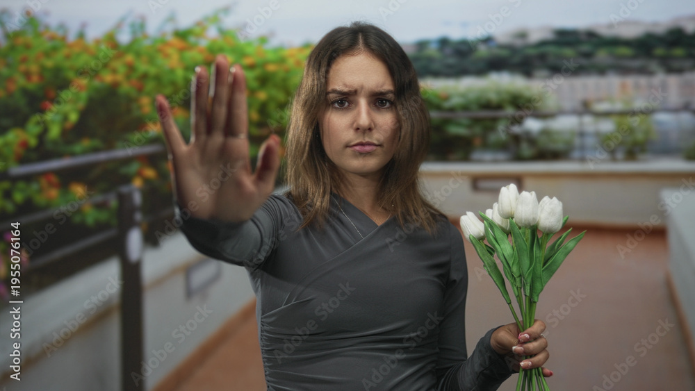 Obraz premium Woman holding white tulips on building terrace raises open hand palm toward camera in a clear stop gesture, frowning slightly; firm refusal.