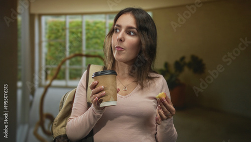Woman holding coffee cup and pastry morsel with hand, wearing backpack and pink sweater inside a building lobby; quiet contemplation.