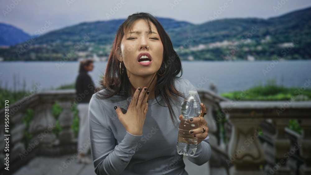Fototapeta premium Young woman holding water bottle, hand at throat, coughing on a building terrace by a lake; discomfort recovery.