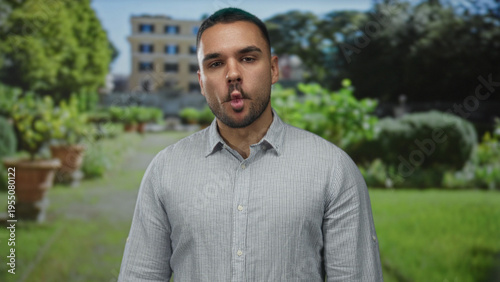 Young hispanic man makes fish face gesture in front of historic building amidst green outdoors; humor joy playfulness spontaneity.