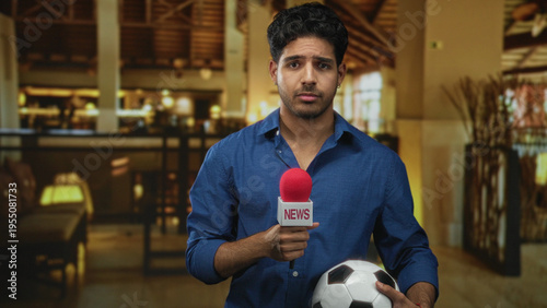 Obraz na plátně Man holds news microphone and cradles soccer ball while making a hesitant gesture in a building lobby; hesitation reporting