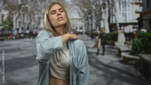 Woman holding neck in pain on busy city street with blurred pedestrians, benches, and bare trees; discomfort.