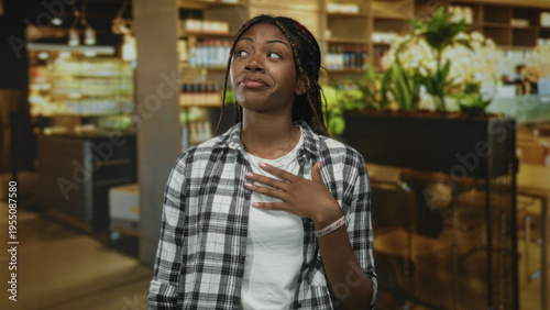 African american woman in flannel shirt with hand on chest, open mouth and braided hair standing in a building restaurant interior near plants and shelves; surprise sincerity apology.
