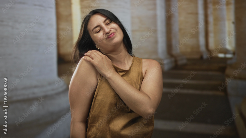 Naklejka premium Woman holding shoulder and wincing in front of a stone building colonnade wearing brown tanktop; discomfort.