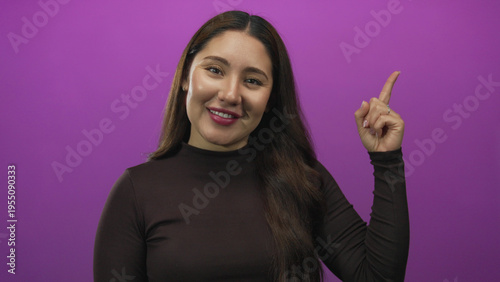 Fotografia Woman points her finger upward in purple studio with smiling expression and upward gaze; inspiration